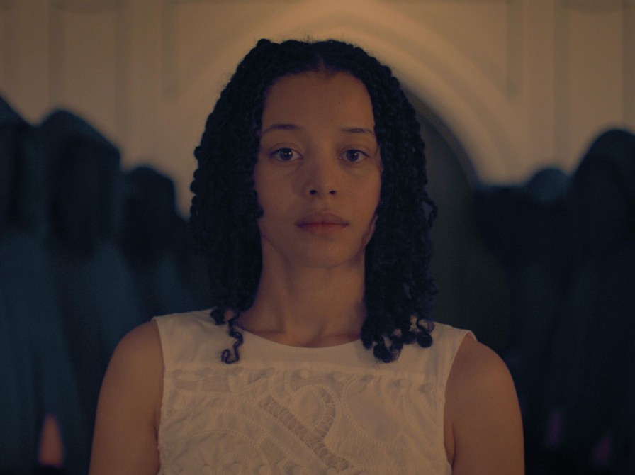 Kvinde i hvid kjole i et indendørsmiljø med mørke former i baggrunden.